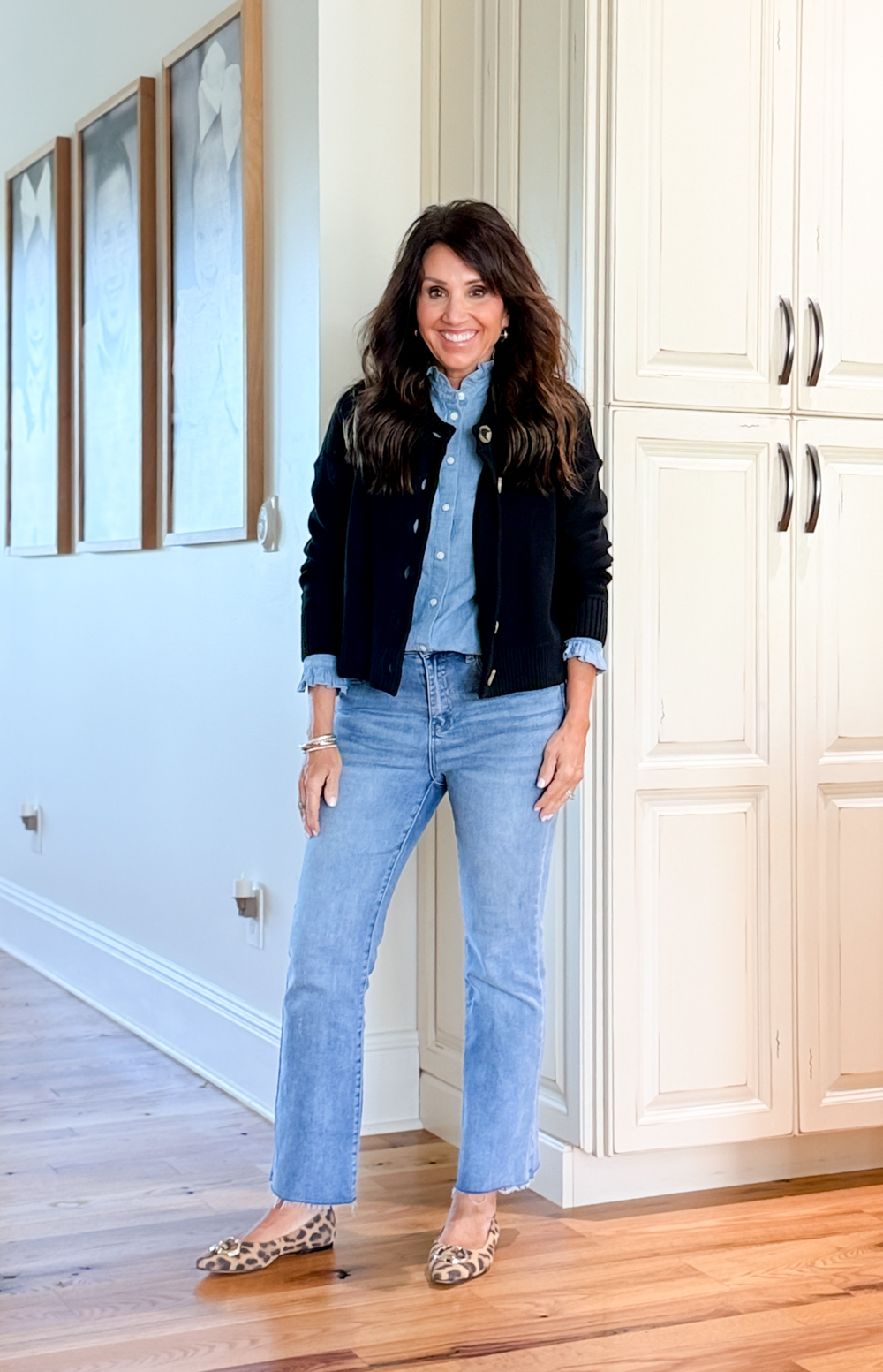 Cyndi Spivey wearing light wash straight leg jeans, chambray shirt, navy jacket, and leopard flats standing in a hallway.