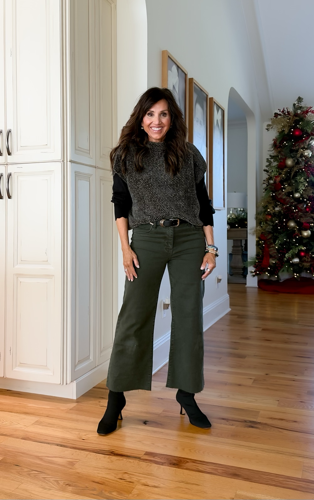 Woman wearing a gray sweater vest over a black top with olive cropped pants and black heeled boots, a casual Thanksgiving outfit idea.