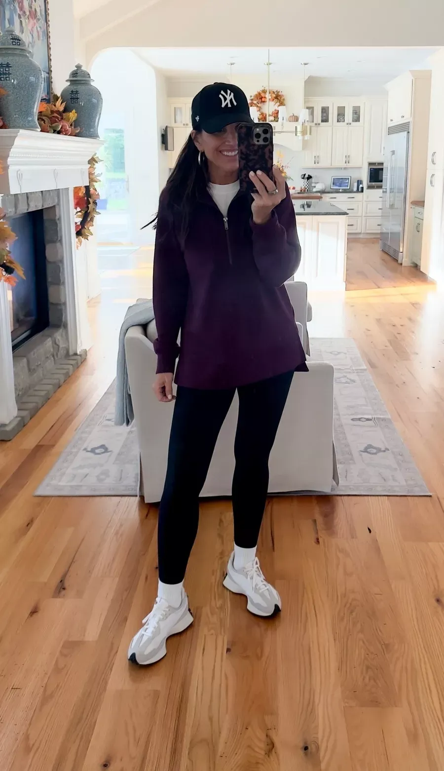 Woman wearing a burgundy half-zip pullover, black leggings, white sneakers, and a black baseball cap, a comfortable Thanksgiving travel or lounging outfit.