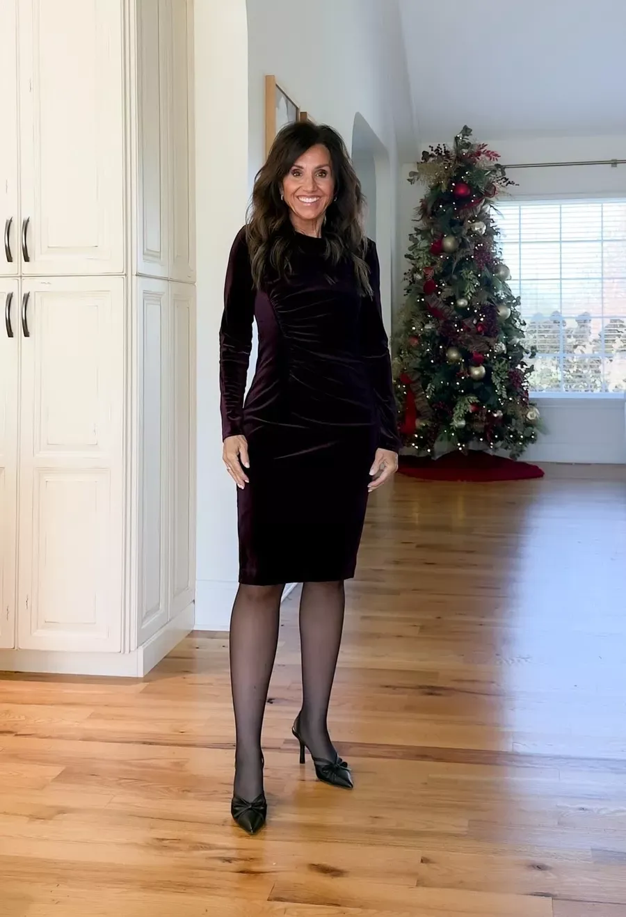 Cyndi Spivey wearing a dark velvet long-sleeve dress with black sheer tights and black heels, standing in a home with a decorated Christmas tree.