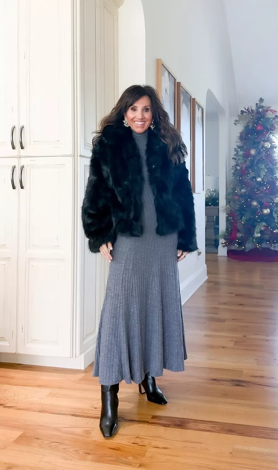 Cyndi Spivey wearing a gray ribbed knit dress with a black faux fur jacket, black heeled boots, and statement earrings in a home with a decorated Christmas tree.