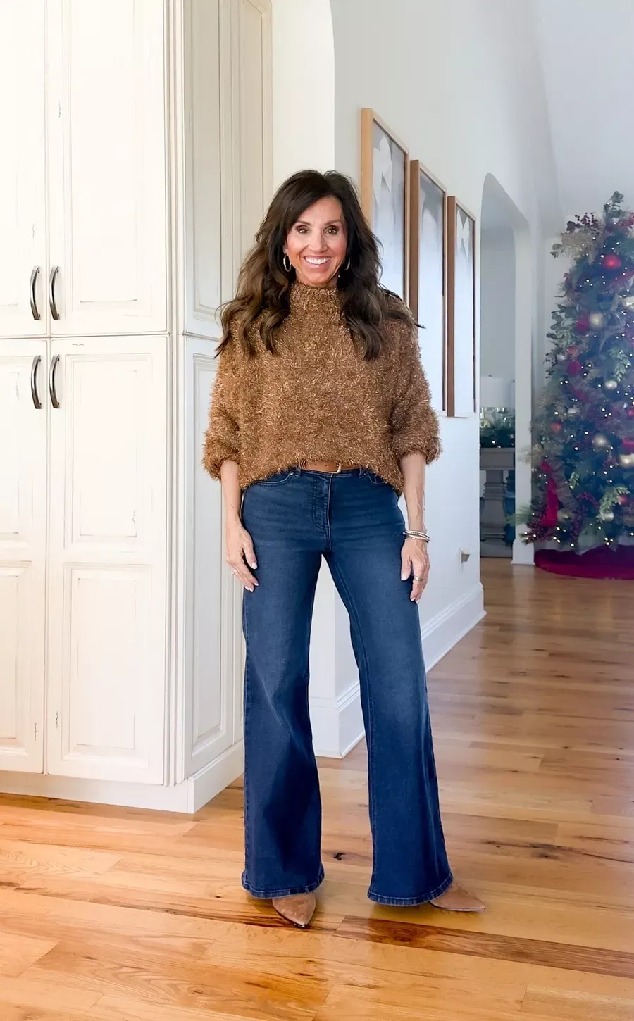 Cyndi Spivey in a brown textured sweater, wide-leg jeans, and tan booties standing in a home with a Christmas tree in the background.