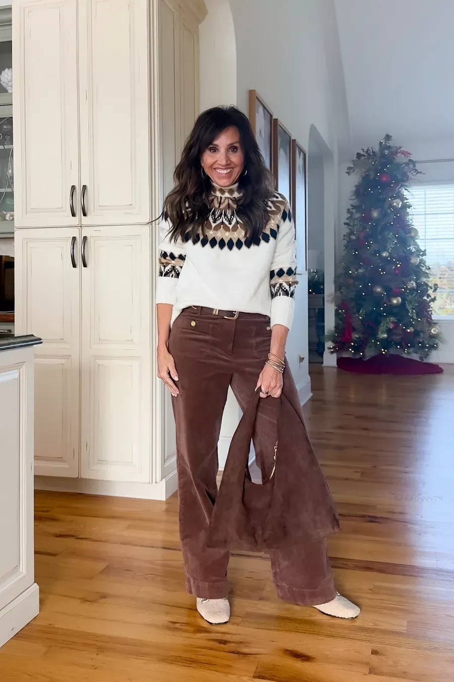 Cyndi Spivey wearing a neutral fair isle sweater with brown corduroy pants and light textured shoes, holding a brown suede bag in a home decorated for Christmas.