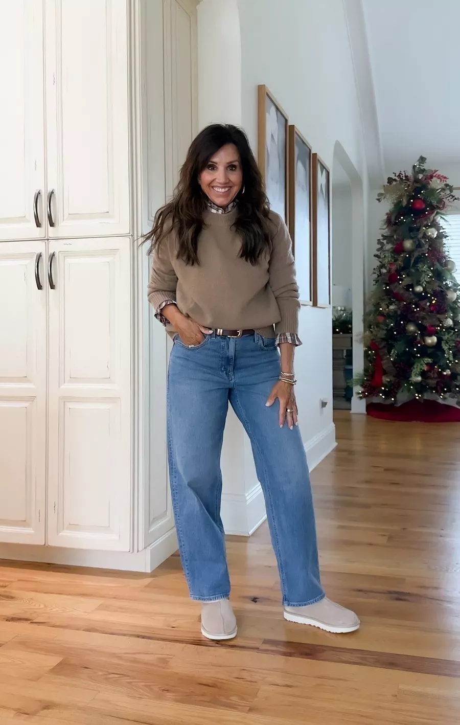 Cyndi Spivey wearing a tan sweater layered over a plaid shirt with light-wash jeans and beige slip-on shoes, standing in a home with a decorated Christmas tree.