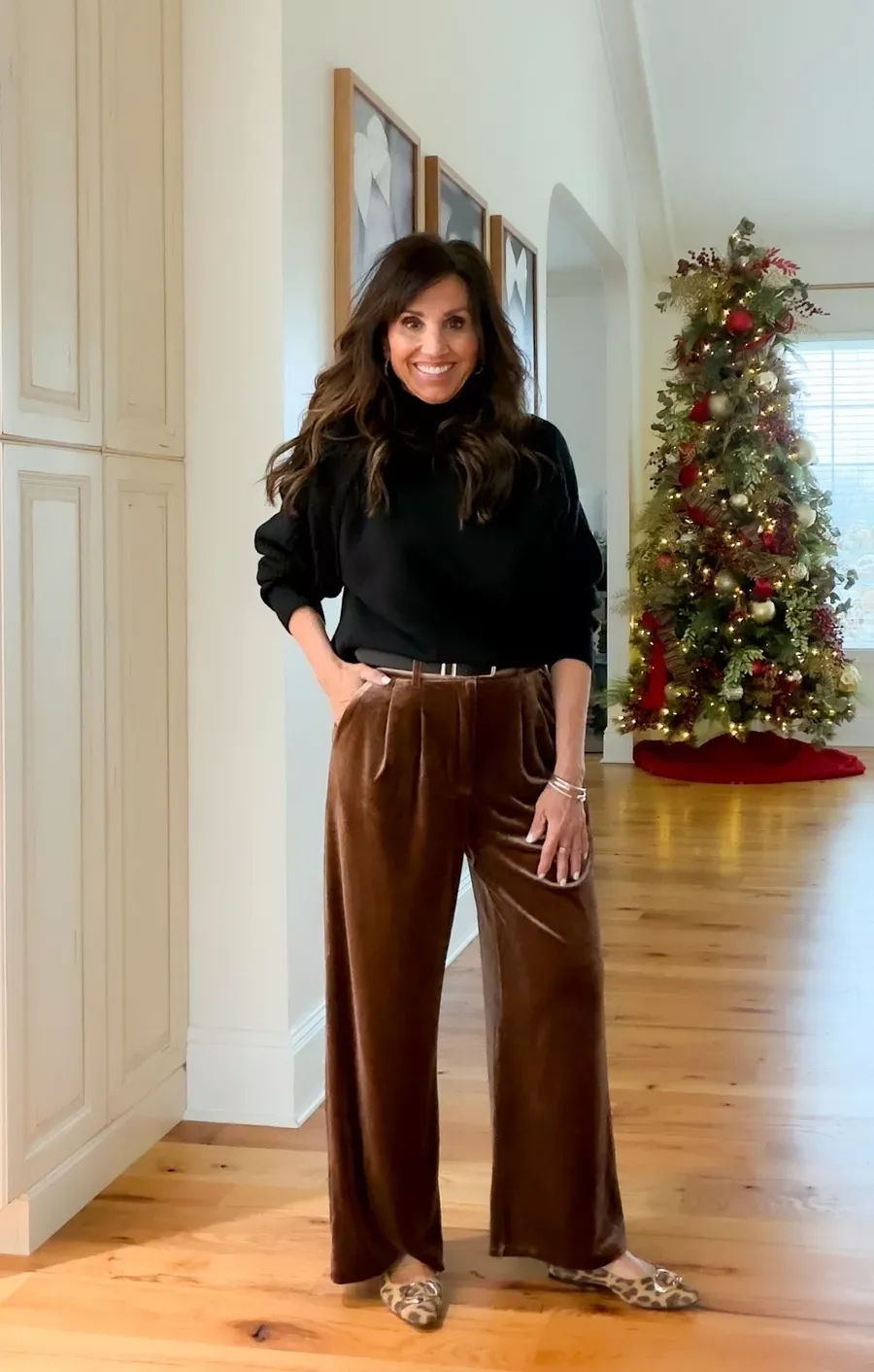 Cyndi Spivey wearing a black sweater tucked into brown velvet wide-leg pants with leopard flats, standing in a decorated hallway with a Christmas tree behind her.
