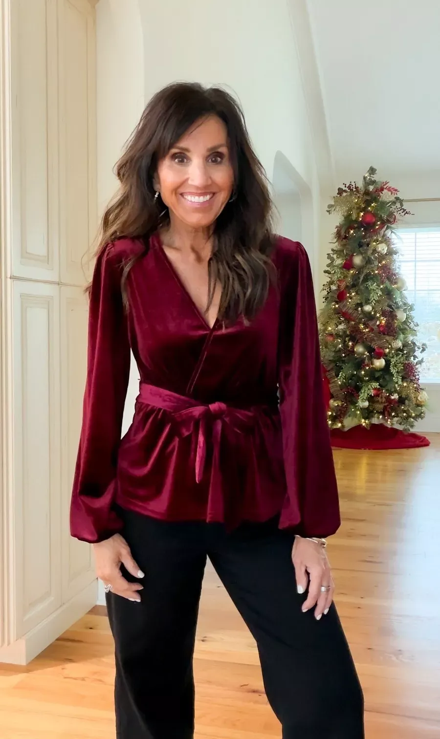 Cyndi Spivey wearing a deep red velvet wrap top with a tie waist and black pants, standing indoors near a Christmas tree decorated with red and gold ornaments.