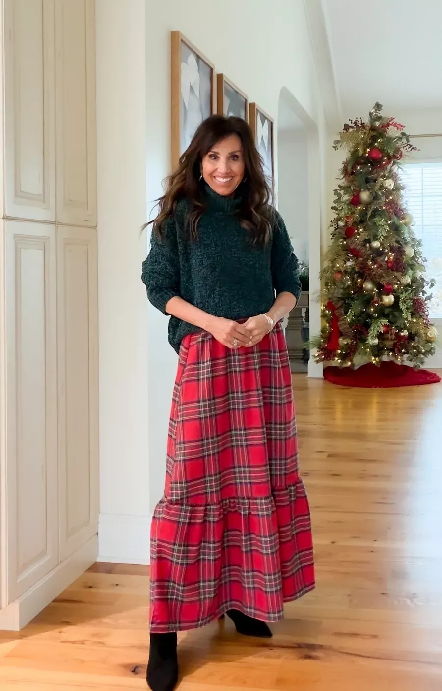 Cyndi Spivey wearing a green turtleneck sweater with a red plaid maxi skirt and black boots, standing indoors with a decorated Christmas tree in the background
