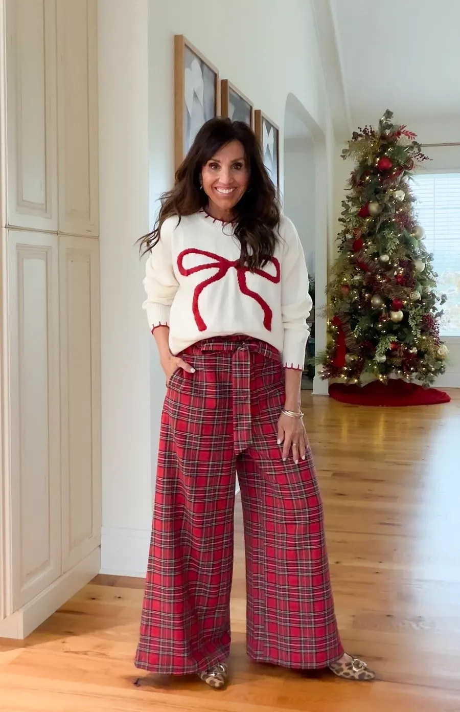 Cyndi Spivey wearing a white sweater with a red bow motif and red plaid wide-leg pants, standing indoors with a decorated Christmas tree in the background