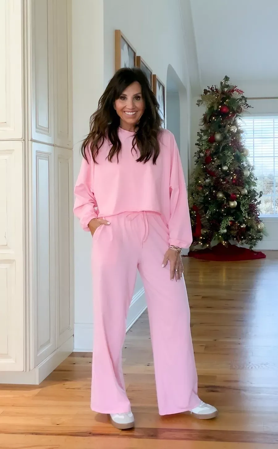 Cyndi Spivey wearing a colorful light pink matching lounge set with wide-leg pants and white sneakers, standing in a decorated living room with a Christmas tree