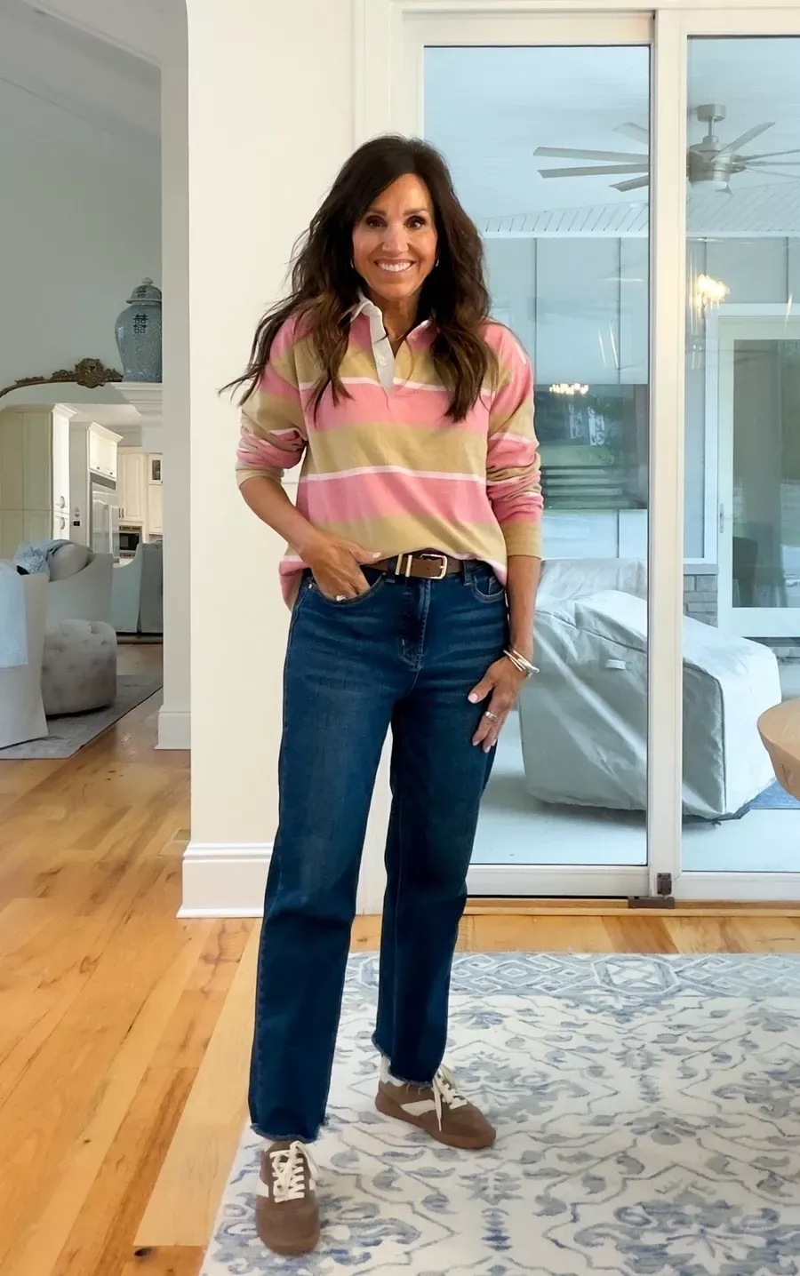 Cyndi Spivey wearing a pink and tan striped rugby pullover with dark wash jeans and brown sneakers, standing in a bright room with wood floors and a large window