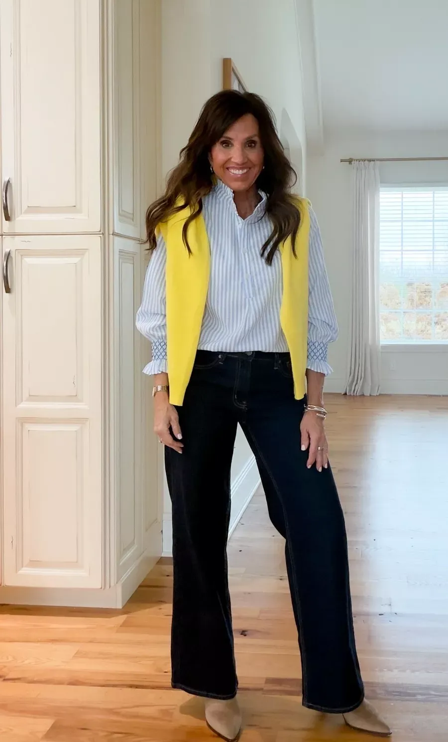 Cyndi Spivey wearing a light blue striped blouse with a yellow sweater draped over her shoulders, paired with dark wash wide-leg jeans and tan boots, standing in a kitchen with wood floors