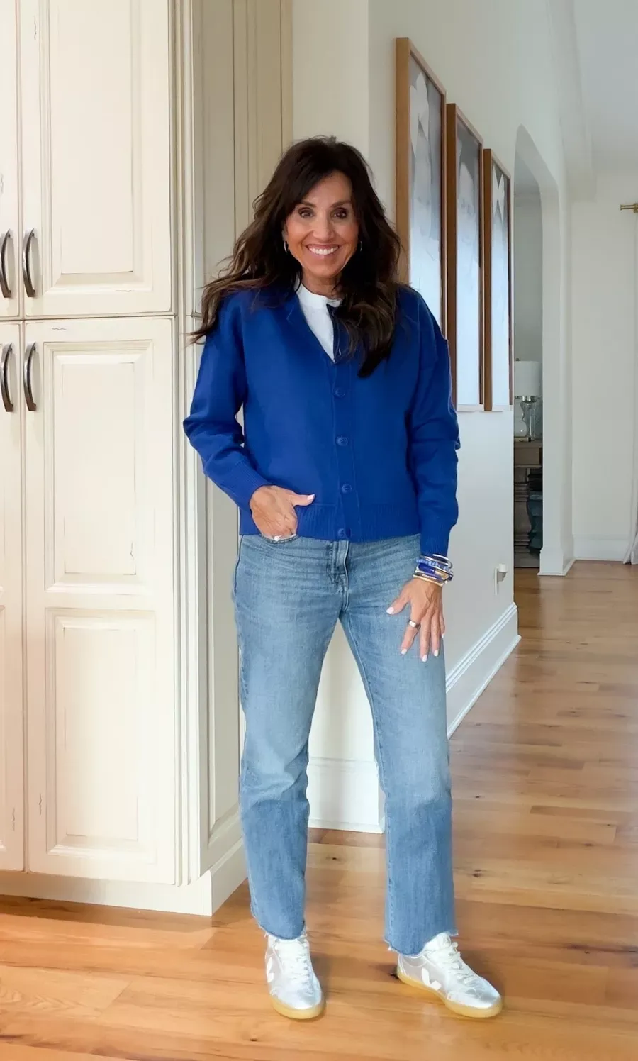 Cyndi Spivey wearing a colorful cobalt blue cardigan over a white t-shirt with light wash straight-leg jeans and white sneakers, standing in a bright kitchen space